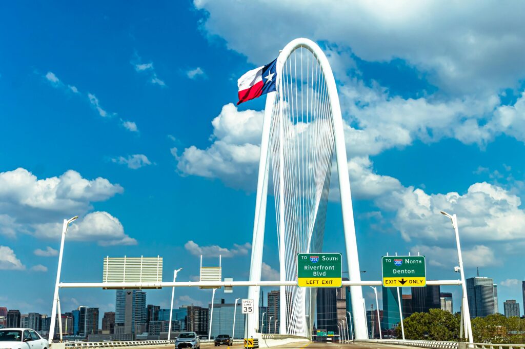 Iconic view of Margaret Hunt Hill Bridge with Texas flag and Dallas skyline under a blue sky.